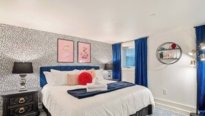 A bedroom with a blue headboard, white bedding, a red pillow, a black nightstand with drawers, a round wall shelf, and a blue curtain. - The Braeburn: Luxury Flat, Steps From NW 23rd! (Portland)