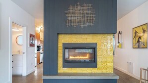 A modern living room with a fireplace, a geometric wall art piece above it, and a kitchen in the background.