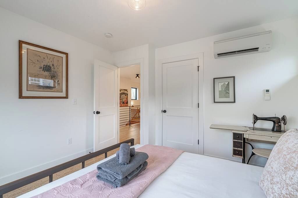 Un dormitorio con una cama, un escritorio con una máquina de coser, un cuadro enmarcado y una puerta que da a otra habitación.