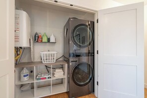 A laundry room with a front-loading washer and dryer, shelves with cleaning supplies, and a white laundry basket. - Bright and modern 2bd/2.5ba- close to Alberta st! (Portland)
