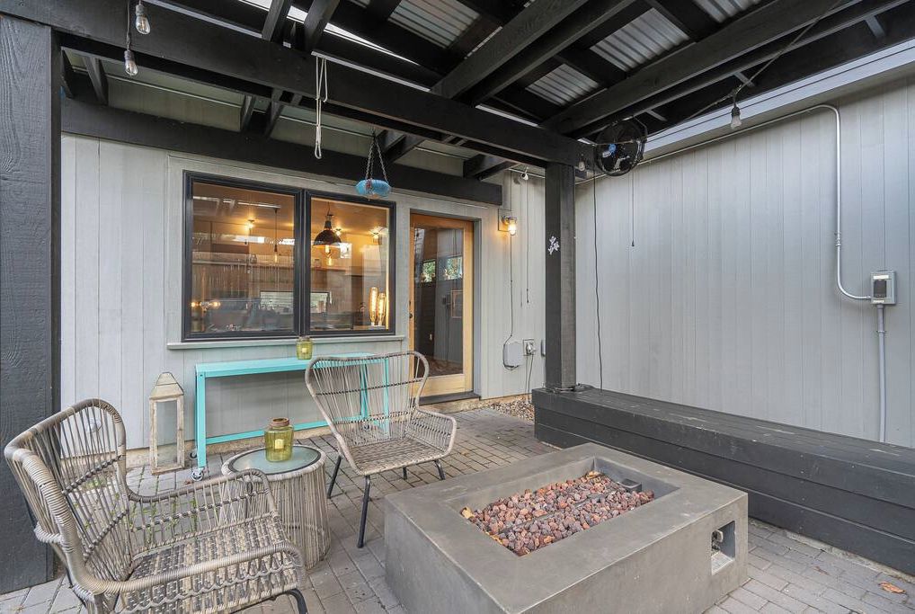 An outdoor patio with wicker chairs, a table, a fire pit, and a view of the interior through a glass door.