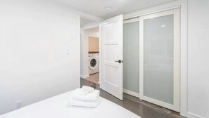 A modern bedroom with a neatly made bed, a white wardrobe, and a visible laundry area in the background.