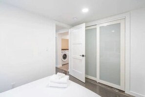 A modern bedroom with a neatly made bed, a white wardrobe, and a visible laundry area in the background.