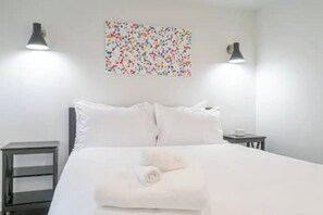 A neatly made bed with white linens and a rolled towel, two bedside tables, and a colorful abstract painting on the wall. - 2 Bed/1 Bath in NE - Close to Mississippi Ave (Portland)