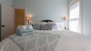 Una cama bien hecha con un edredón estampado, una lámpara de noche y una ventana con cortinas traslúcidas.