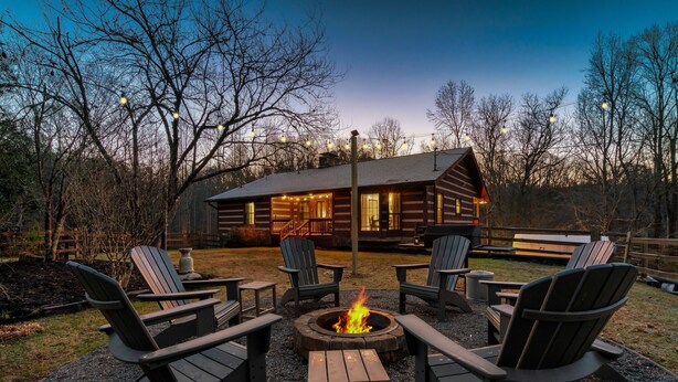 A log cabin with a fire pit surrounded by Adirondack chairs, set against a backdrop of trees and a wooden fence.
