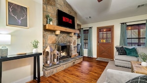 A cozy living room with a stone fireplace, a flat-screen TV mounted above it, and a comfortable seating area with a sofa and a coffee table.