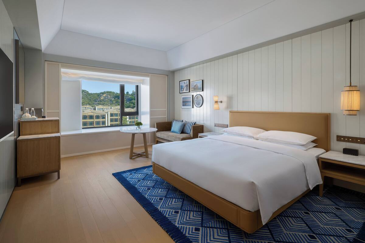Una habitación de hotel moderna con una cama grande, una pequeña mesa, un sofá y vistas a la ciudad a través de un amplio ventanal.