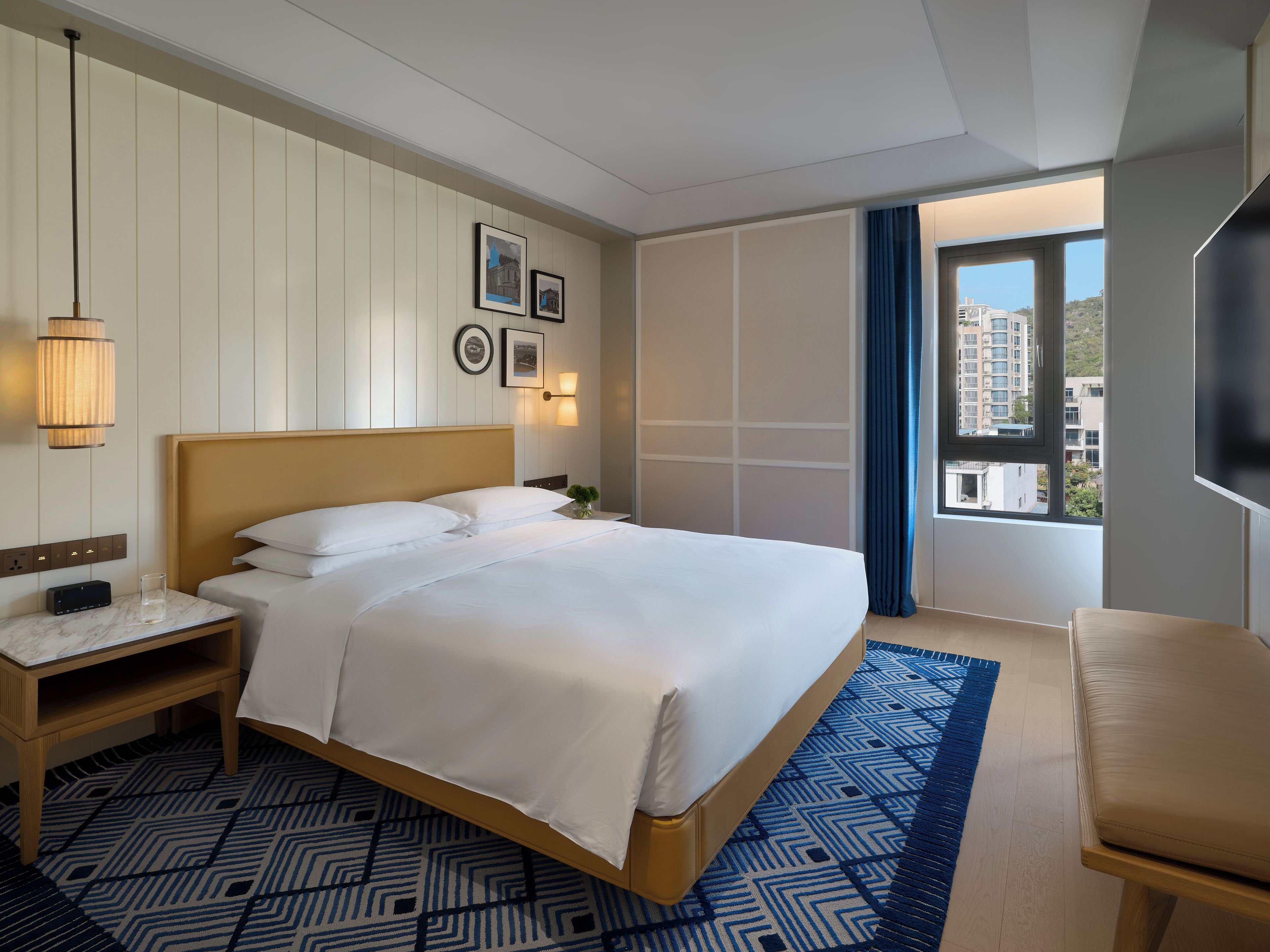 A hotel room with a large bed, bedside table, lamp, and a view of buildings through the window.