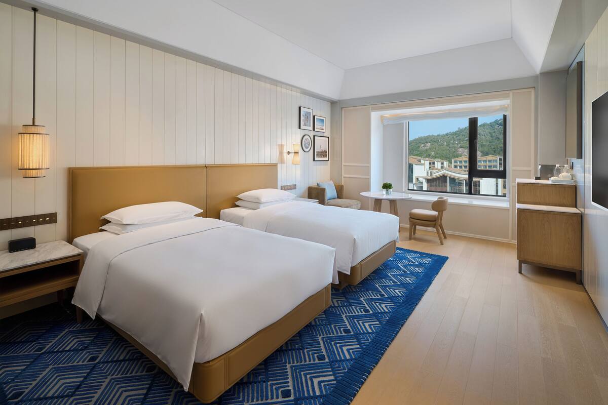Habitación de hotel con dos camas, un escritorio, una silla, un televisor y una ventana con vistas a edificios.