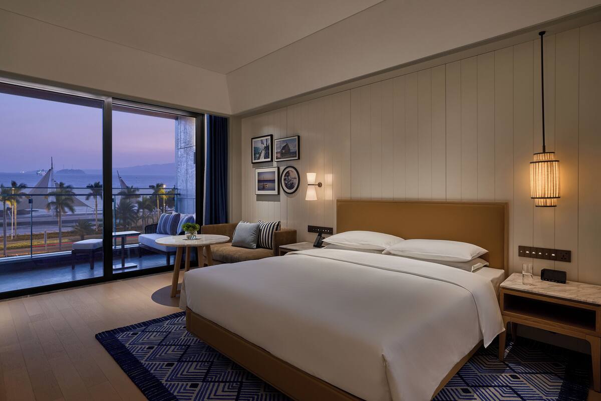 Un hotel moderno con una cama grande, un balcón con vista a la playa y una pequeña zona de estar con una mesa.