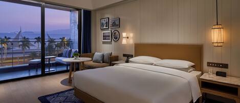 A modern hotel room with a large bed, a balcony with a view of the beach, and a small seating area with a table.