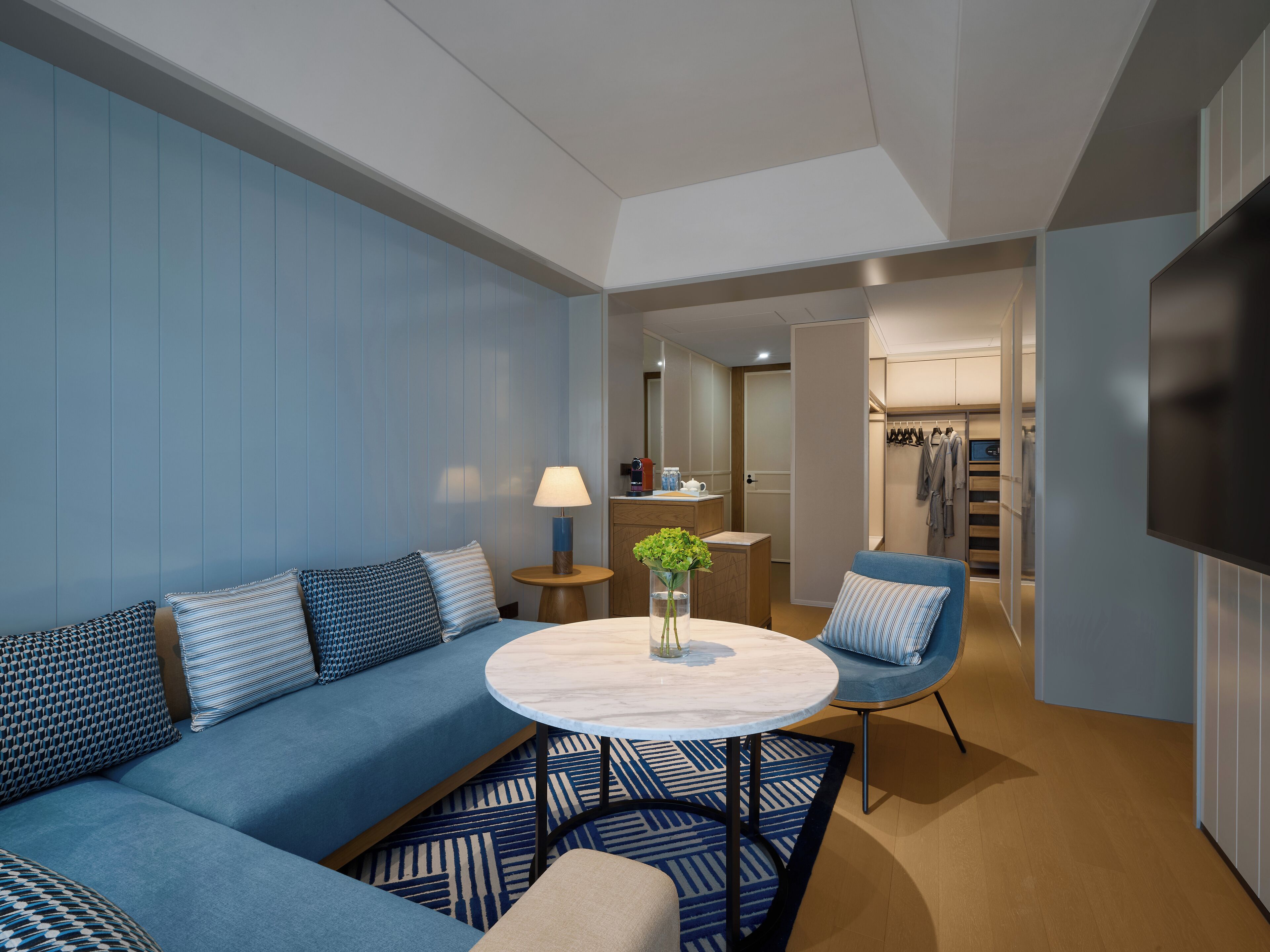 Habitación de hotel moderna con un sofá azul, una mesa redonda, un televisor y un vestidor.