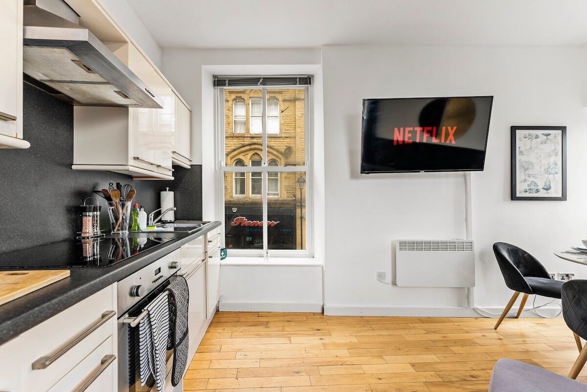 A modern kitchen with white cabinets, a black countertop, and a window with a view of a building. A flat-screen TV mounted on the wall displays the Netflix logo.