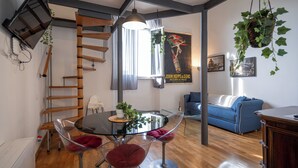 Ein zeitgemäßer Wohnraum mit einem runden Glastisch, transparenten Stühlen, einem blauen Sofa, einer Holzleiter und einer hängenden Pflanze.