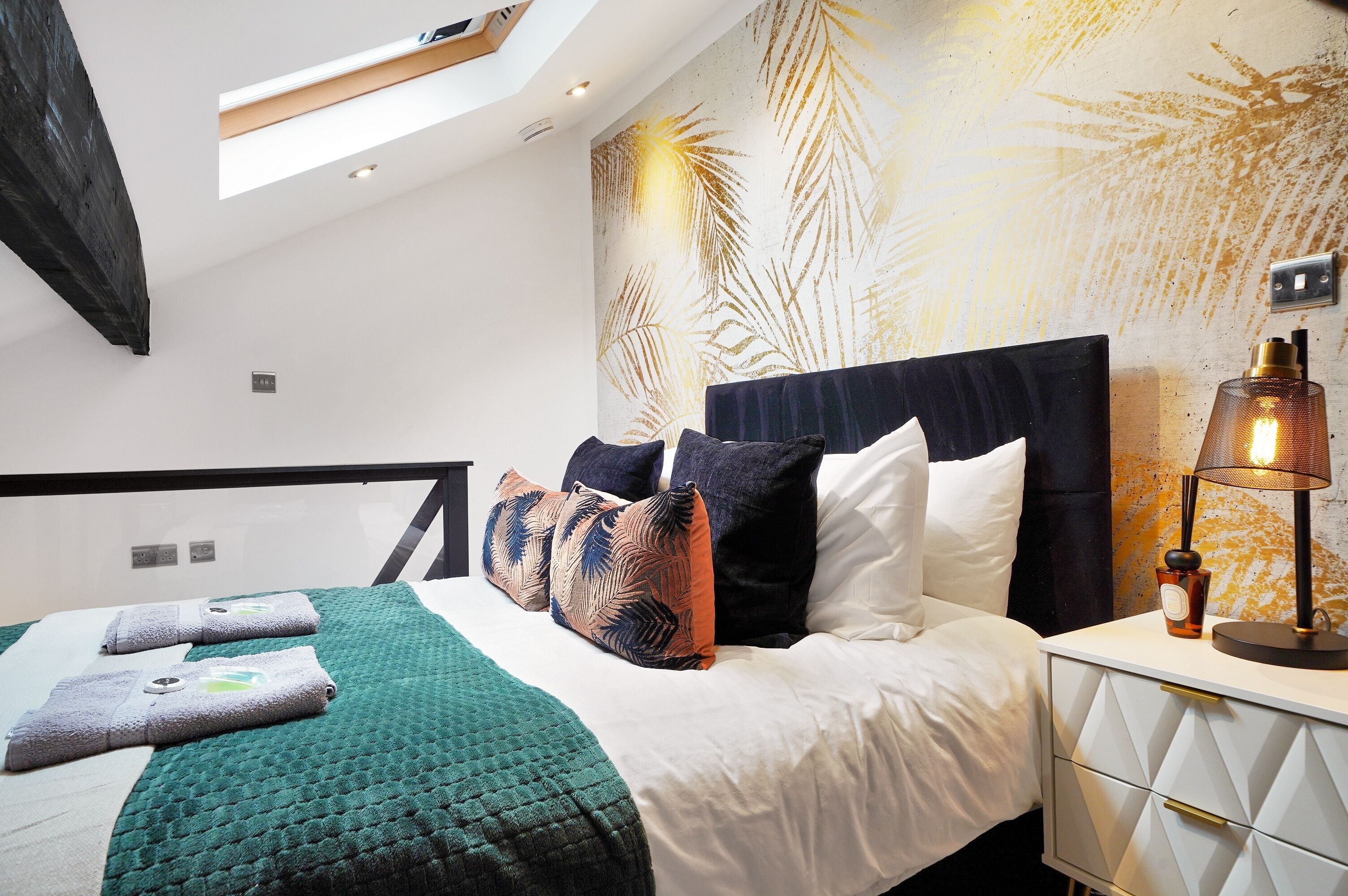 A modern hotel room with a skylight, a bed with a green quilt, multiple pillows, and a bedside table with a lamp.