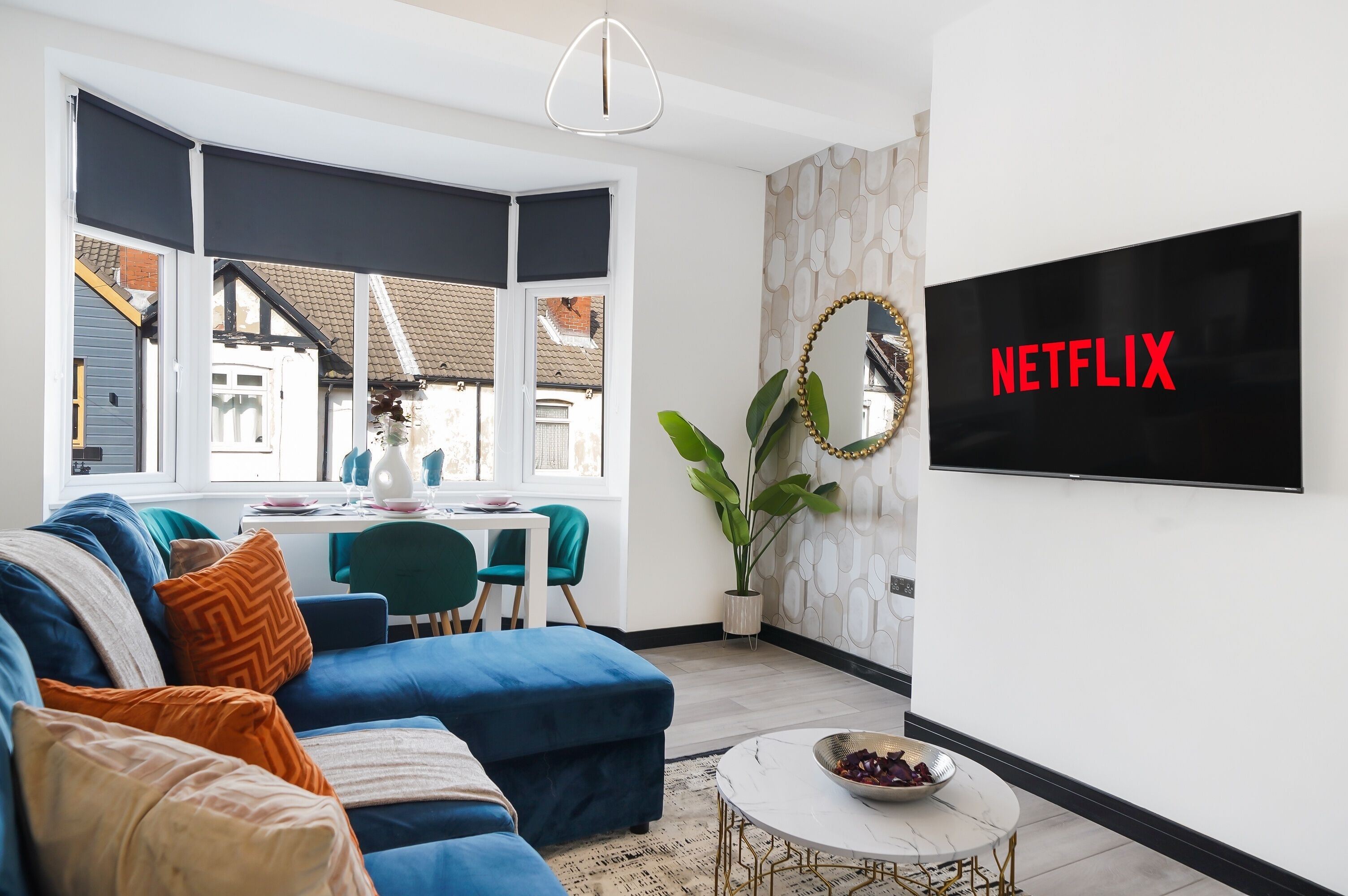 Moderni olohuone, jossa on sininen sohva, ruokailutila pöytineen ja tuoleineen, suuri ikkuna ja seinälle asennettu televisio, jonka näytöllä näkyy teksti ”NETFLIX”.