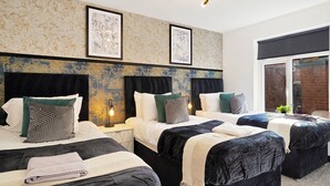 A hotel room with three beds, each with a black headboard, white bedding, and various decorative pillows. There are framed artworks on the wall above the beds, and a modern chandelier hangs from the ceiling. - Elegant Yorkshire Abode - Great Location - Sleeps 19 (Castleford)