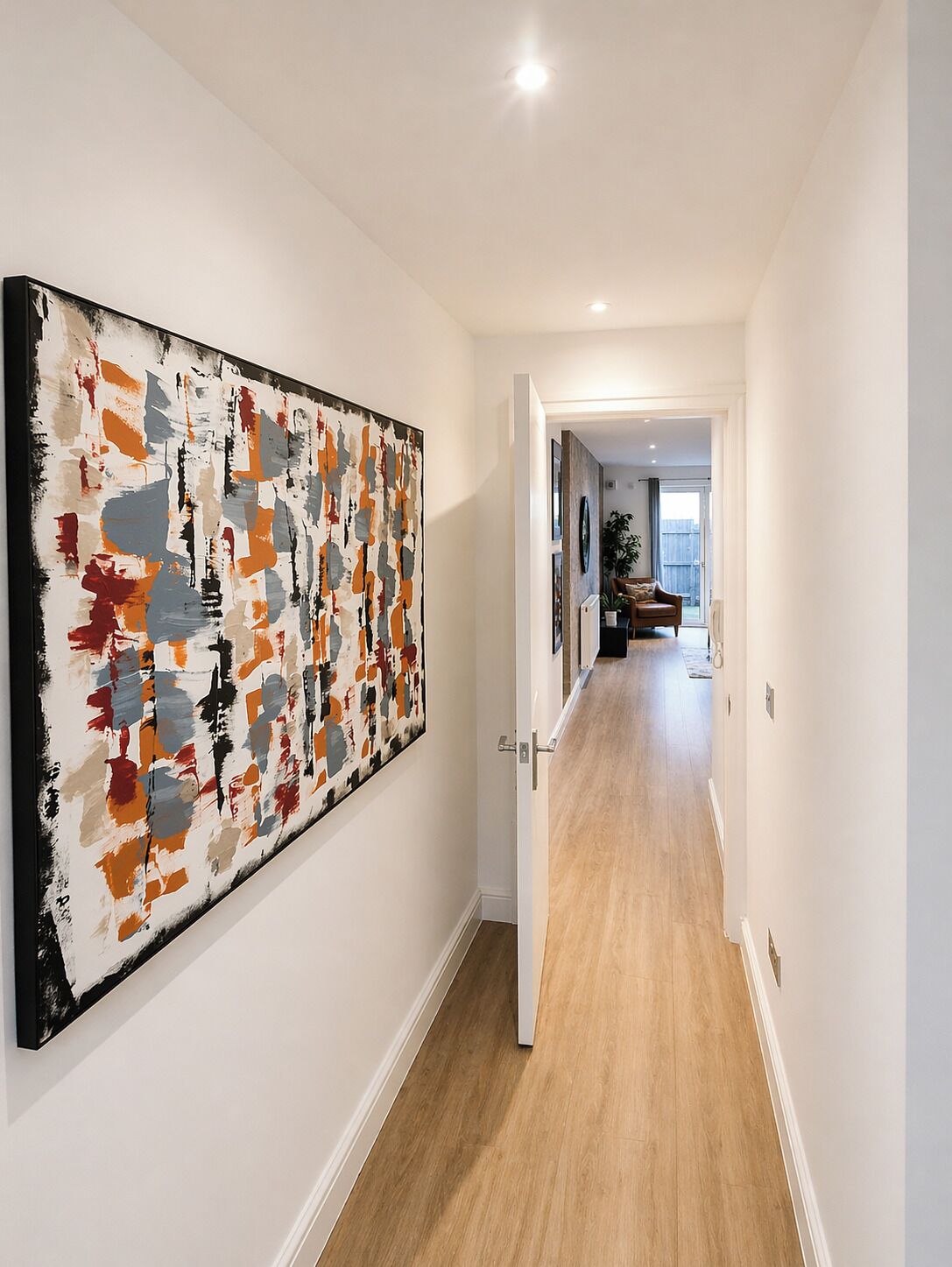 Un pasillo con un cuadro abstracto grande a la izquierda, que conduce a una sala de estar con un sofá y una planta.