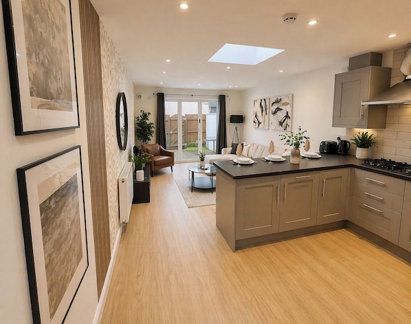 A modern kitchen with a central island, a dining area with a sofa and table, and a view through glass doors to an outdoor space.