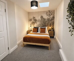 A bedroom with a bed, two bedside tables, a wall mural, a skylight, and a plant.