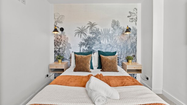 A neatly made bed with a white and orange quilt, two decorative pillows, and a rolled towel. There are two bedside tables with lamps, and a wall mural depicting tropical foliage. - Spacious Bristol Apartment - Parking & WIFI (Bristol)