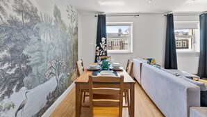 A dining area with a wooden table set for four, a sofa, and a large window with curtains. - Spacious Bristol Apartment - Parking & WIFI (Bristol)