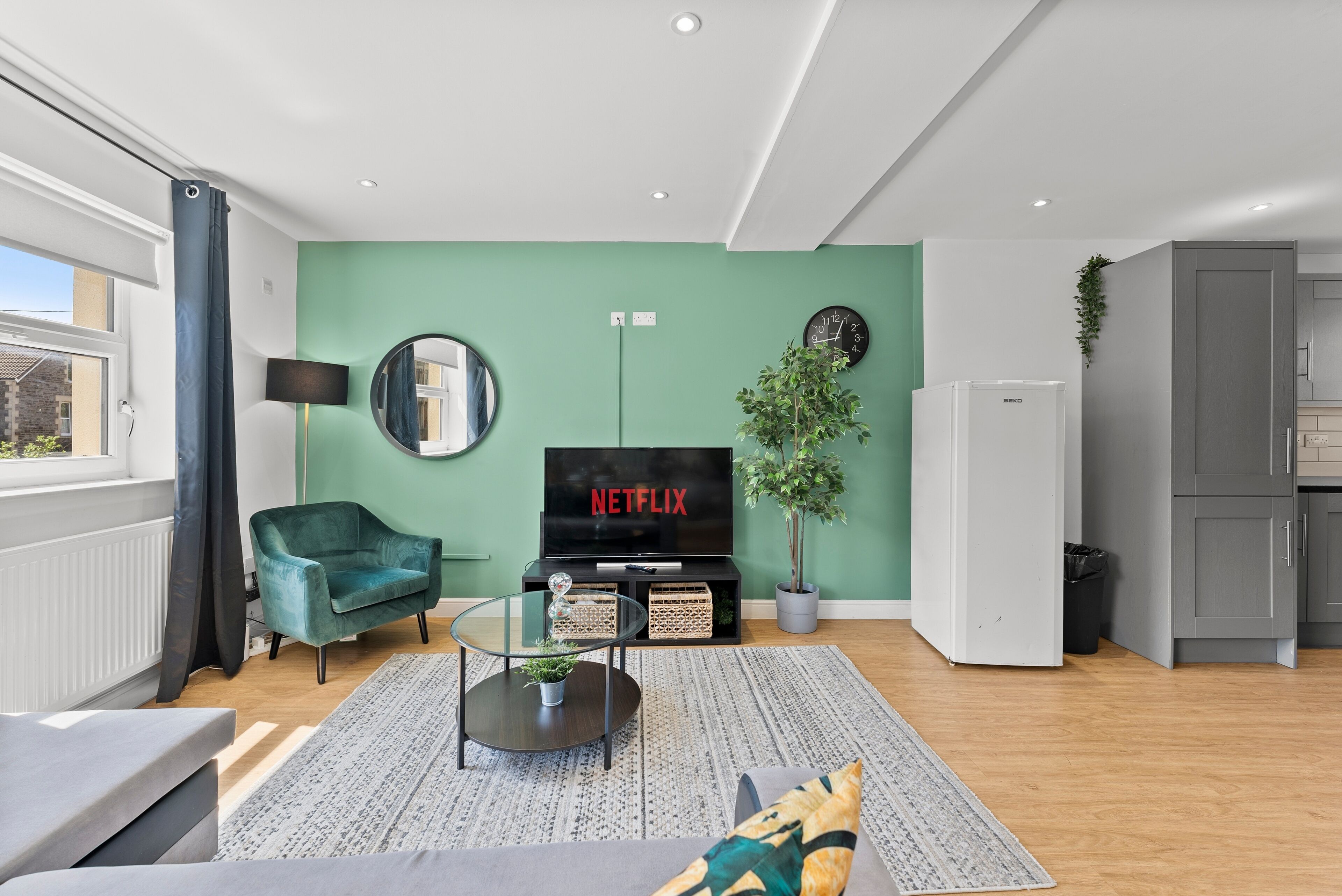 Una sala de estar moderna con una pared de acento verde, un televisor de pantalla plana, un sillón azul verdoso, una mesa de centro de vidrio y un ventanal grande.