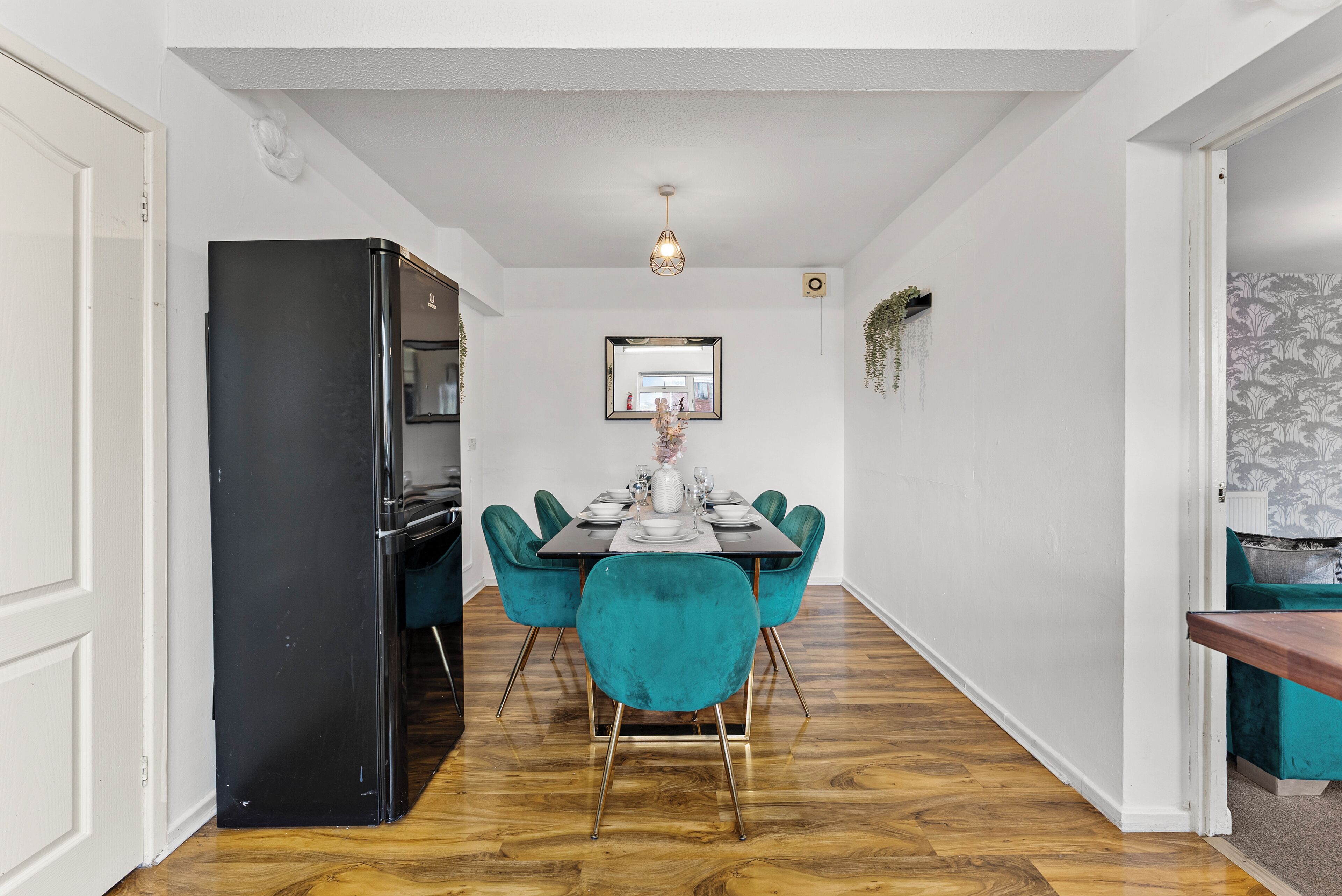 A dining area with a black refrigerator, teal chairs, a wooden dining table, and a hanging light fixture.