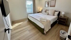 Ein modernes Schlafzimmer mit einem großen Bett, einem Deckenventilator, einem Nachttisch mit Lampe, einem an der Wand montierten Fernseher und einem Fenster mit Jalousien.