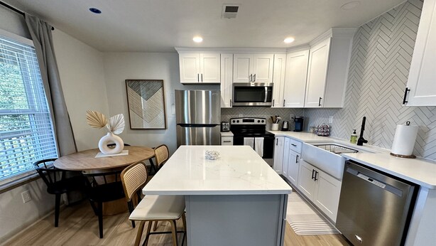 A modern kitchen with white cabinetry, a stainless steel refrigerator, a microwave, and a dishwasher. There is a dining area with a table and chairs, and a window with blinds. - 2 King Beds + 400ft from Beach - 8 (norfolk)