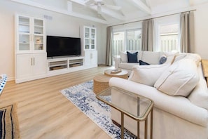 A modern living room with a flat-screen TV, white sofas, a glass coffee table, and a large window with curtains.