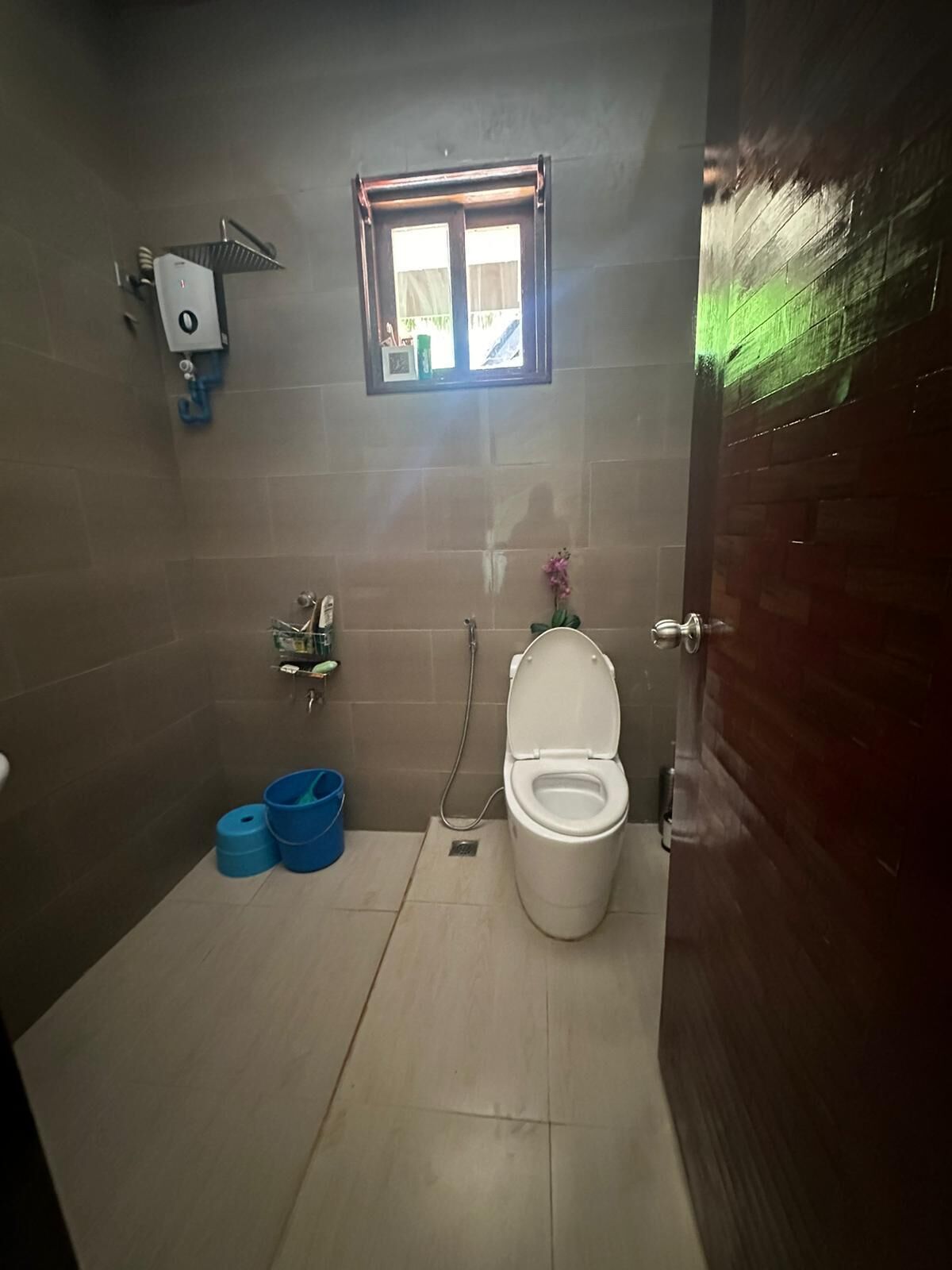 Een kleine badkamer met toilet, douche en een raam. Op de vloer staan twee blauwe emmers.