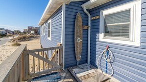Ein Haus direkt am Strand mit einer Holzterrasse, einem Surfbrett, das an der Wand lehnt, und einem Schlauch, der an der Seite hängt.