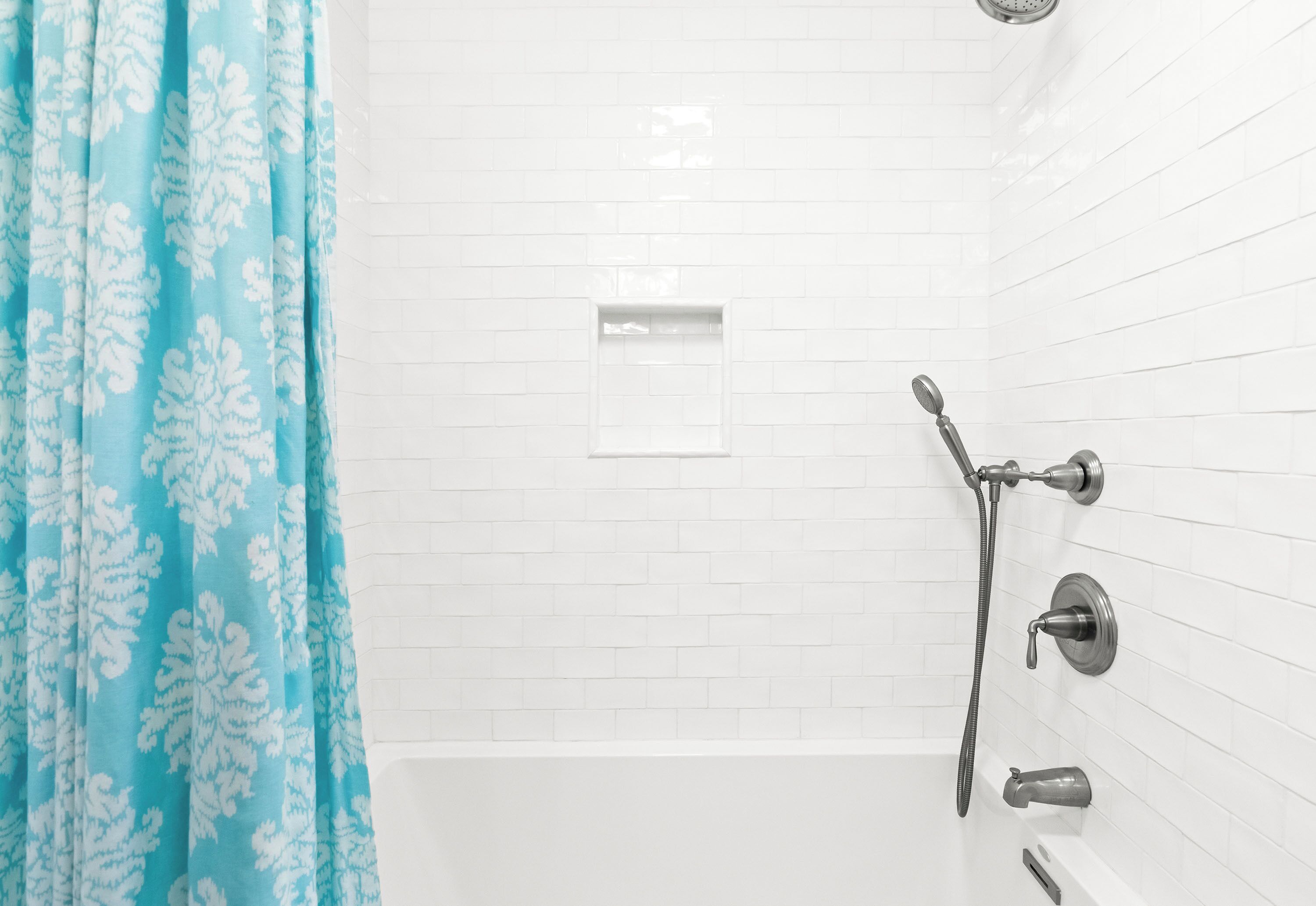 A bathroom with white tiled walls, a shower with a handheld showerhead, and a blue patterned shower curtain.