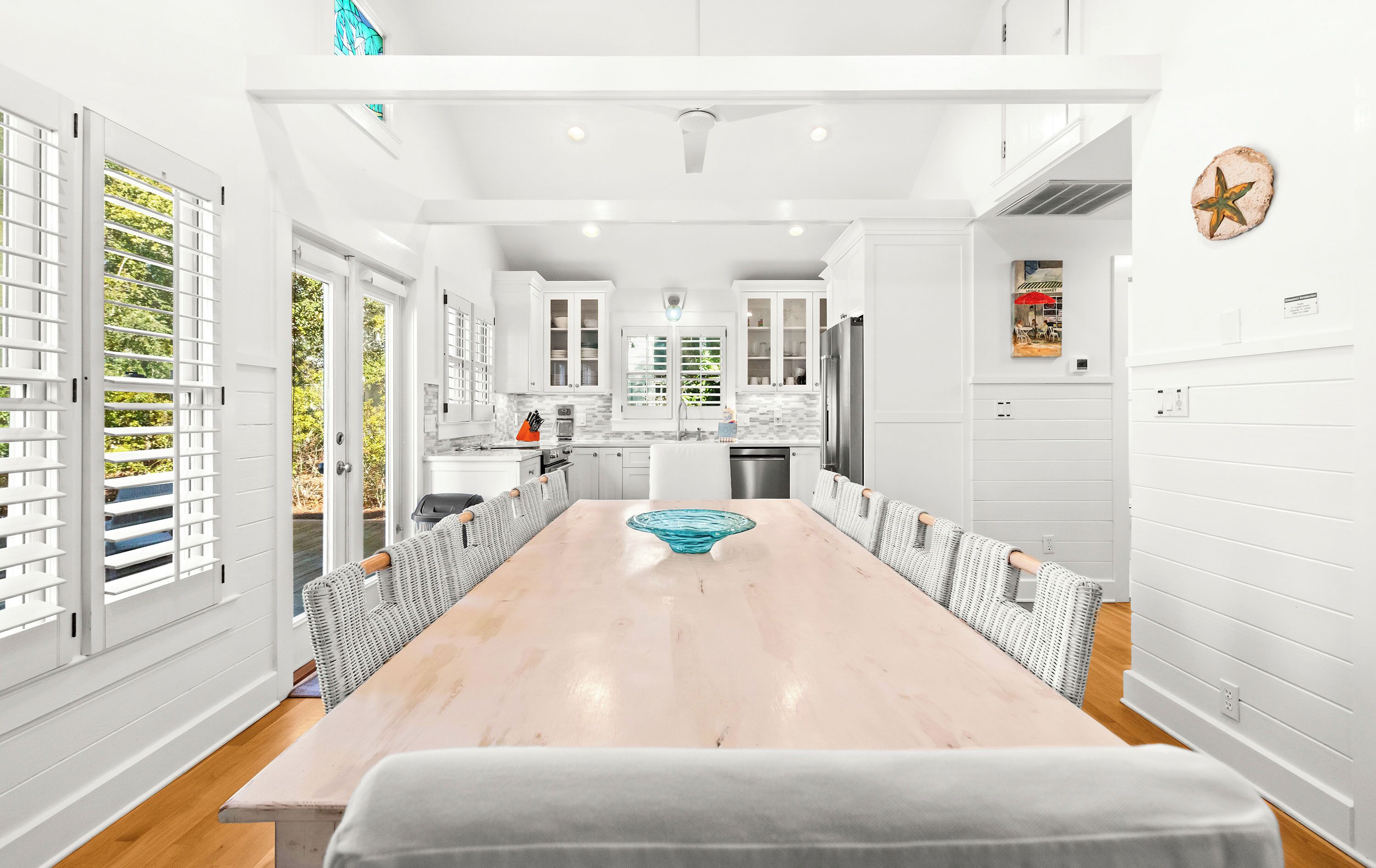 A modern kitchen with white cabinets, a central island, and a dining area with a long table and wicker chairs.