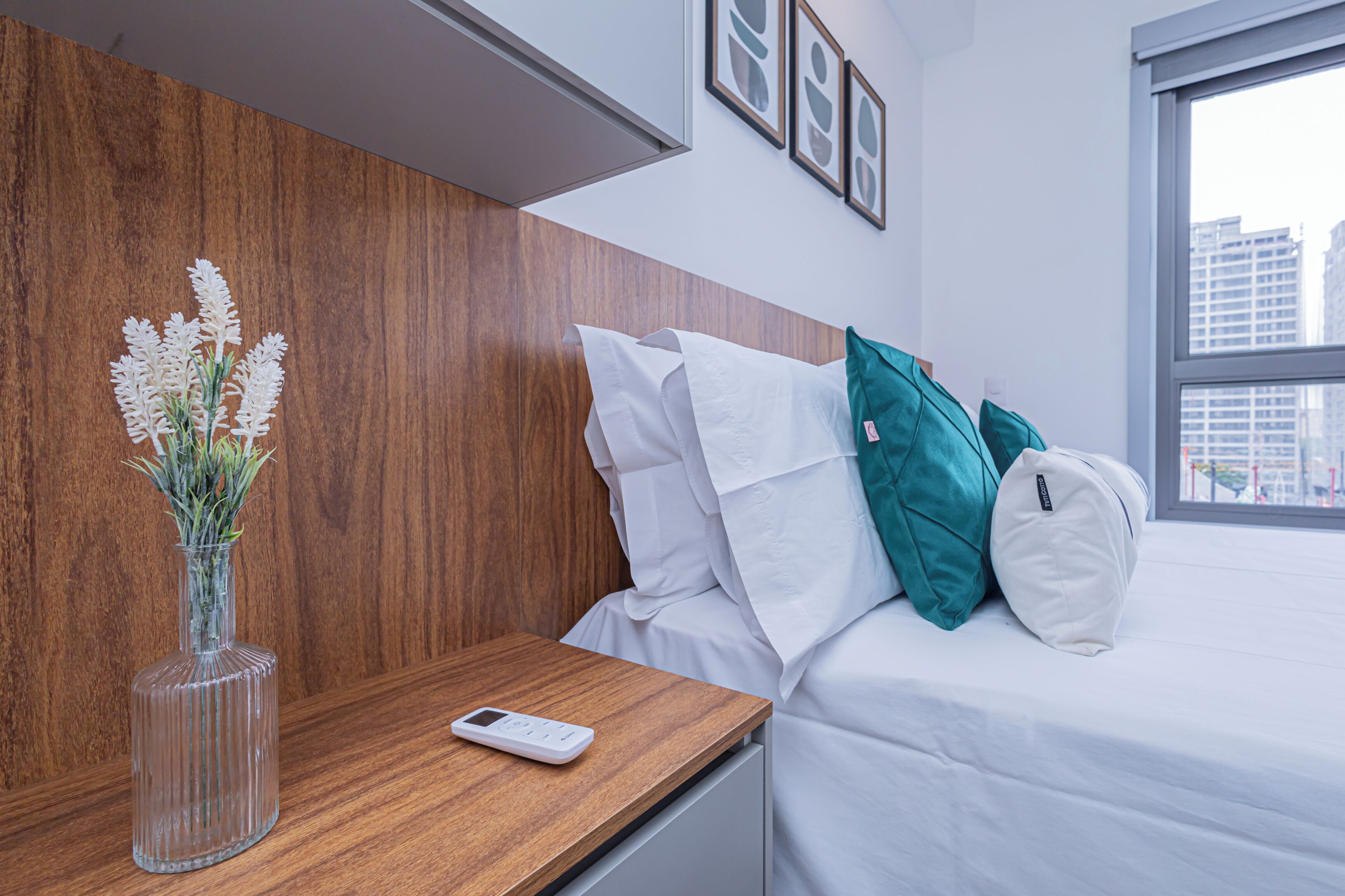 Habitación de hotel con cabecera de madera, ropa de cama blanca, almohada verde azulada y un jarrón transparente con flores blancas sobre una mesita de noche de madera.