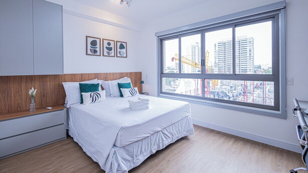 Une chambre d’hôtel moderne avec une grande fenêtre, un lit bien fait, une tête de lit en bois et une petite table de chevet agrémentée d’un vase de fleurs.