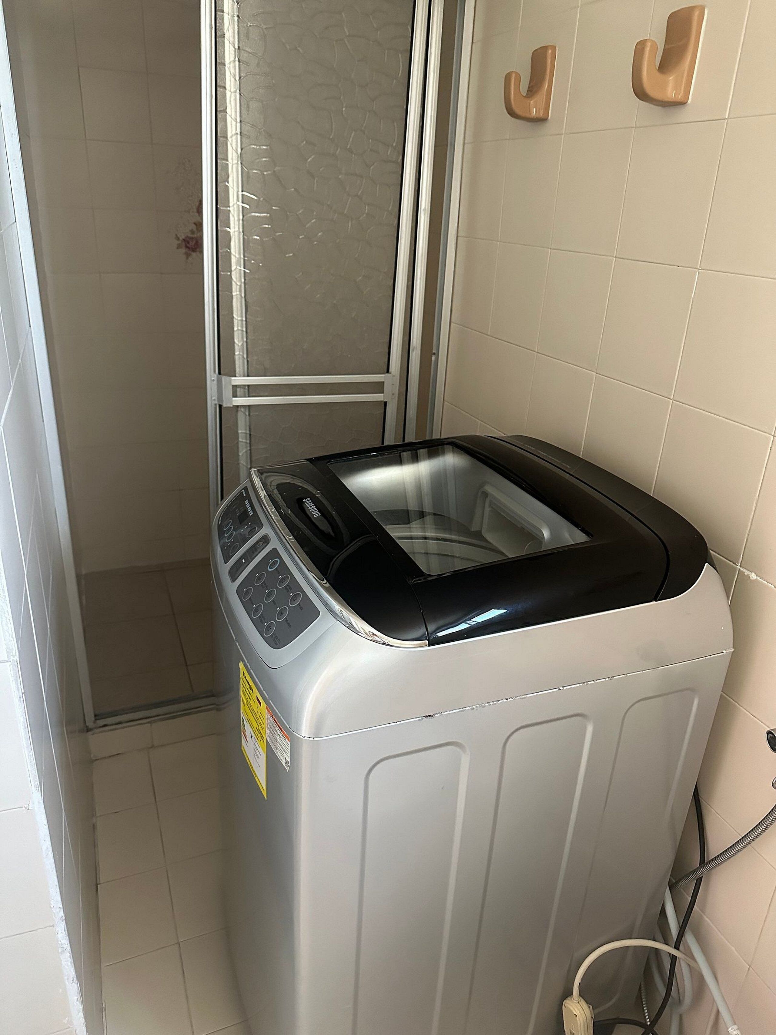 A compact, modern laundry machine in a small, tiled room with a glass door and hooks on the wall.