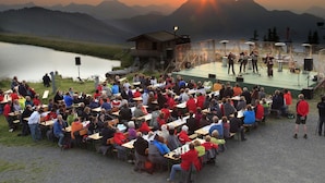 Evento al aire libre con escenario y público sentado en mesas, con vista a un lago y montañas.