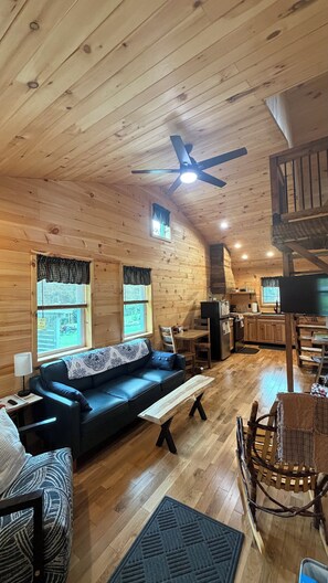 Un interior de cabaña acogedor, con techo de madera, ventilador de techo, sofá, mesa de comedor y una mecedora.