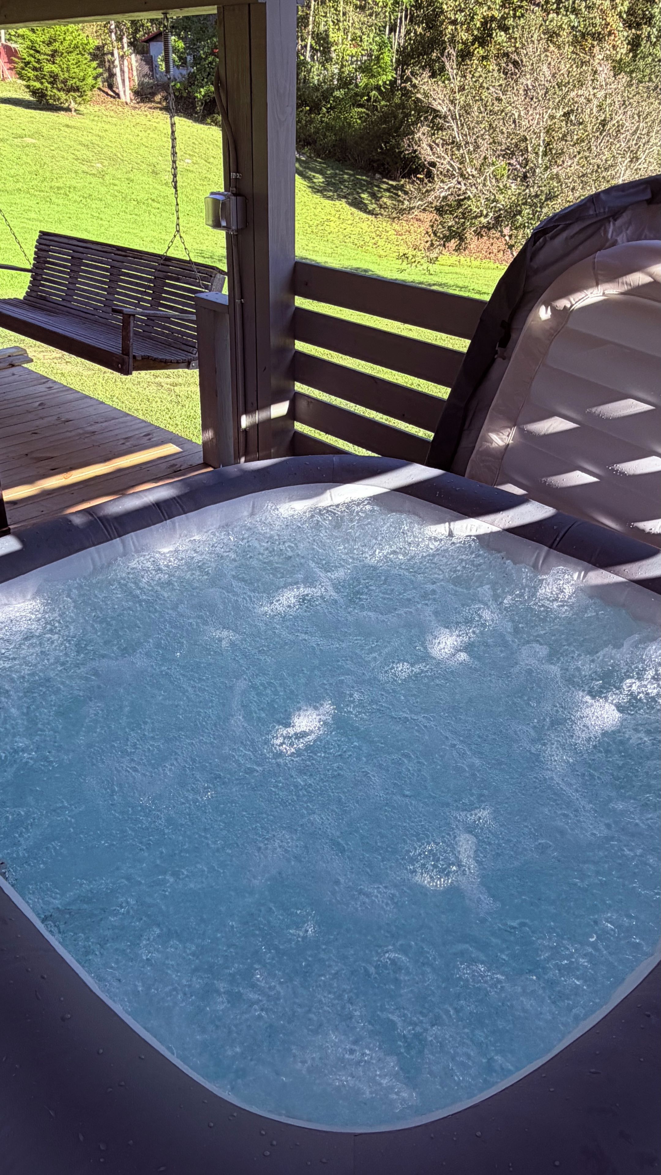Ein Whirlpool mit Holzterrasse und Sitzbank, umgeben von einem Rasen und Bäumen.
