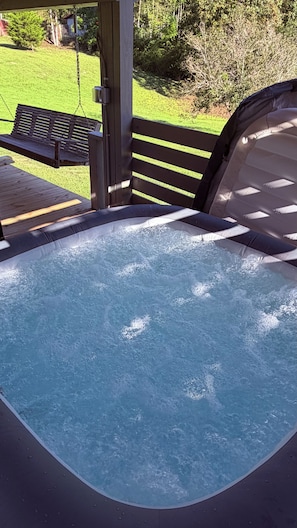 A hot tub with a wooden deck and a bench, surrounded by a grassy yard and trees.