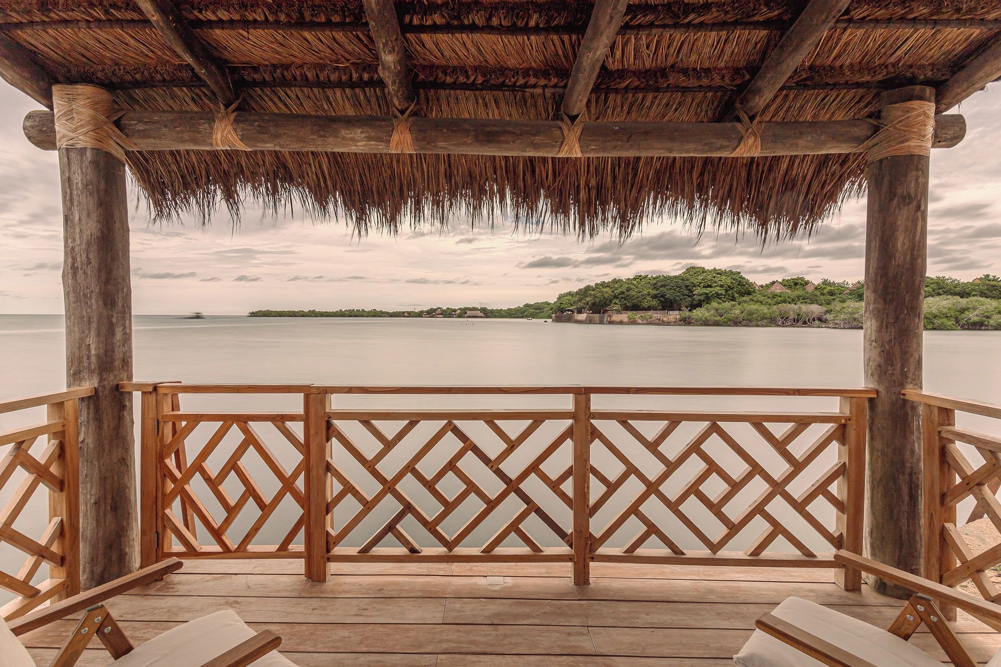 A wooden deck with a thatched roof overlooking a calm body of water and a distant island.