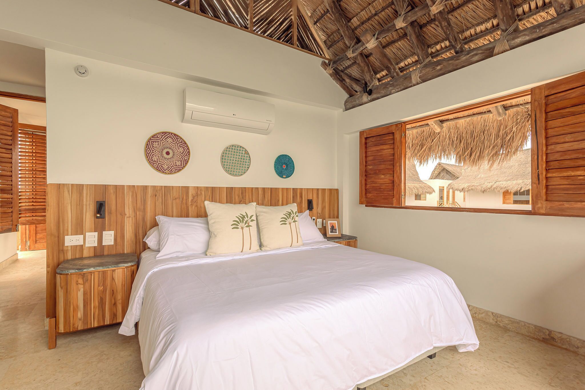 A bedroom with a wooden headboard, a large bed with white bedding, a small bench, and a window with a view of a thatched-roof structure.