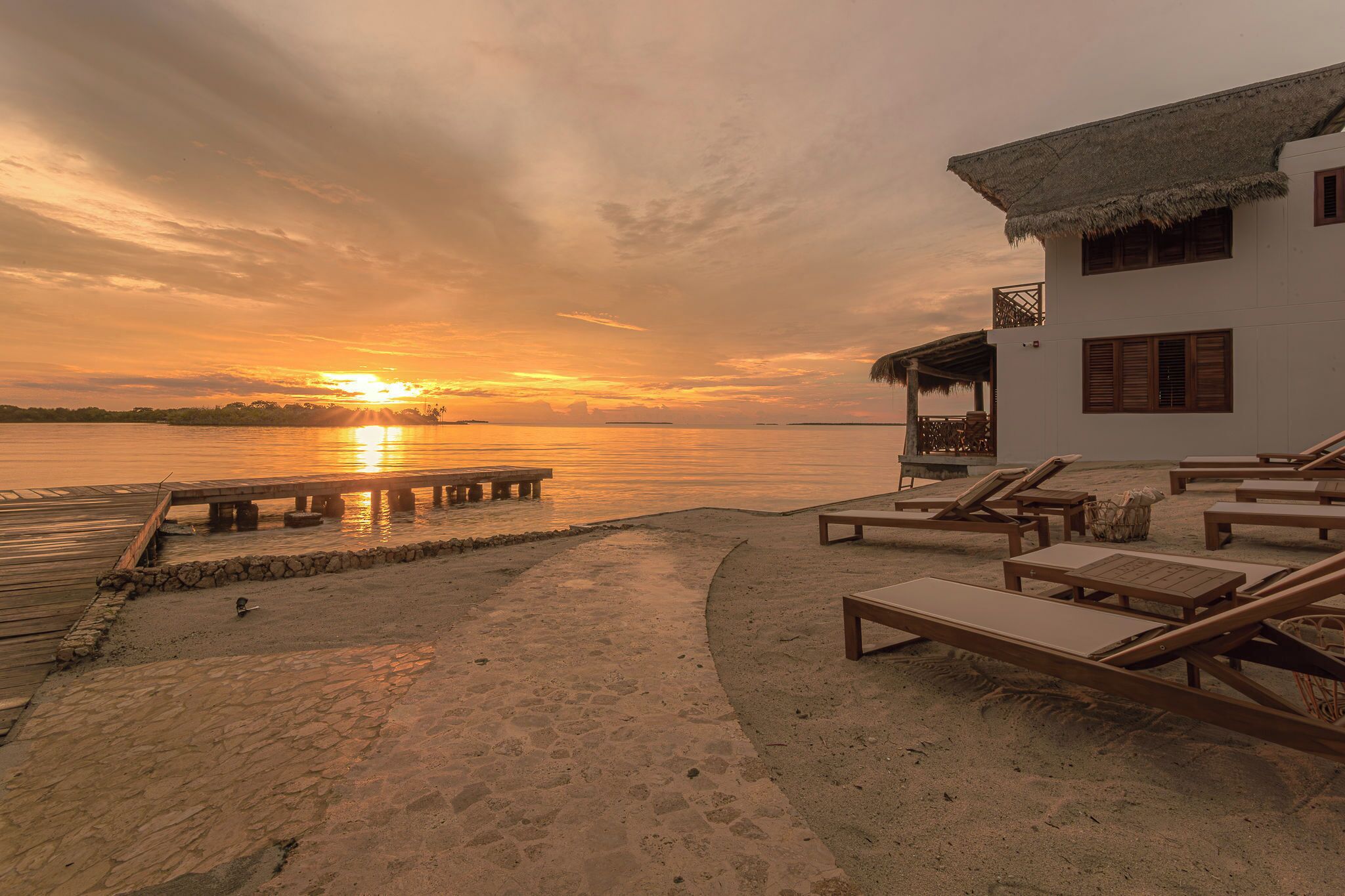 A beachside resort with a wooden deck, lounge chairs, and a building with a thatched roof.