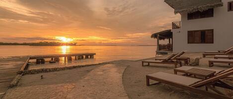 A beachside resort with a wooden deck, lounge chairs, and a building with a thatched roof.