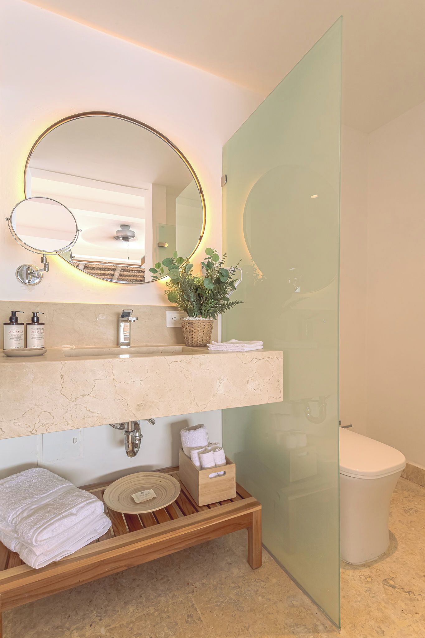 A bathroom with a marble countertop, a round mirror with a gold frame, a potted plant, and a toilet.