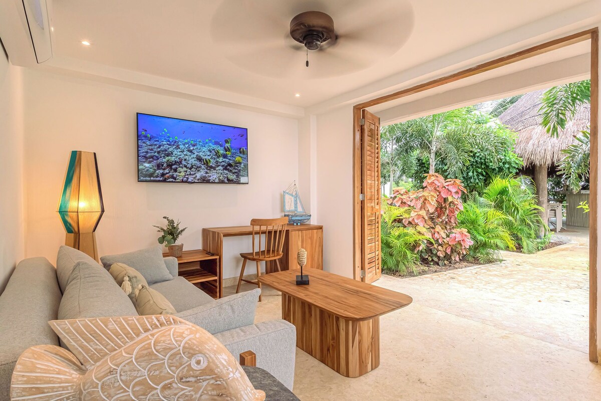 Un salon moderne avec un canapé, un téléviseur, une table en bois et une vue sur un jardin tropical.