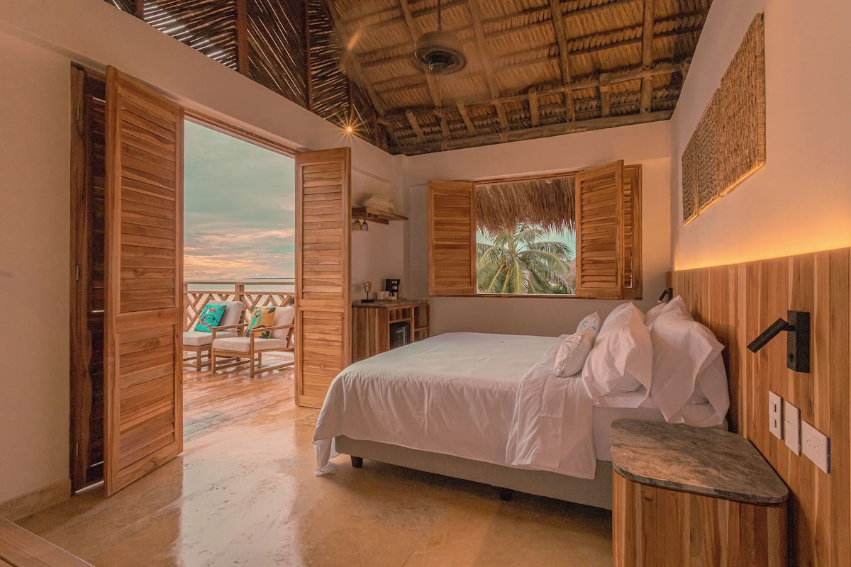 A bedroom with a bed, wooden furniture, a view of palm trees, and a thatched ceiling.